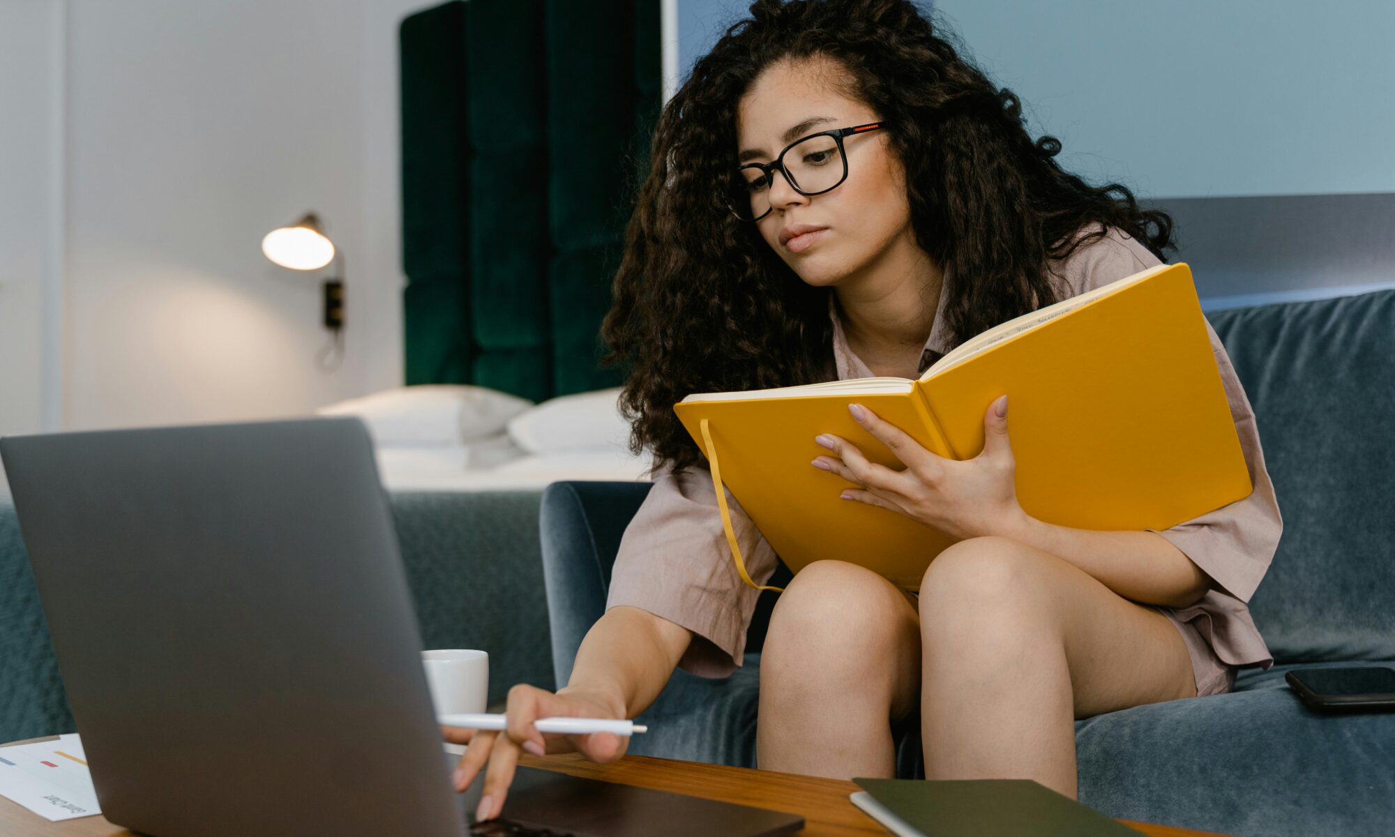 a woman sits in front of her laptop with a book in her hand. she is typing on her laptop and using the book as notebook to make notes