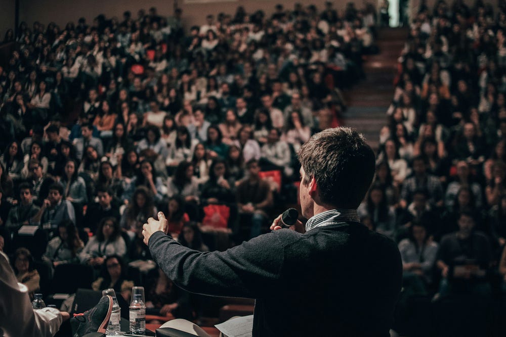 A photograph of a smartly-dressed, white male giving a speech in front of a crowd